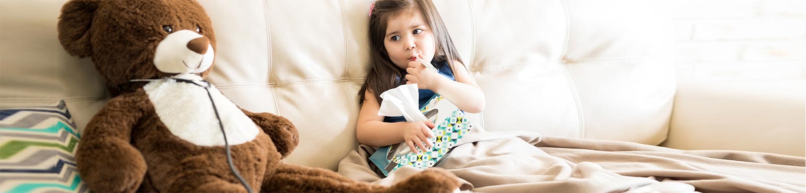 Little girl holding a tissue box sitting next to a teddy bear on a couch