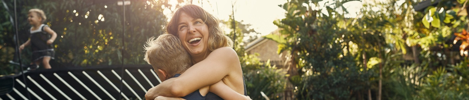 Mother hugging kids in garden next to trampoline 