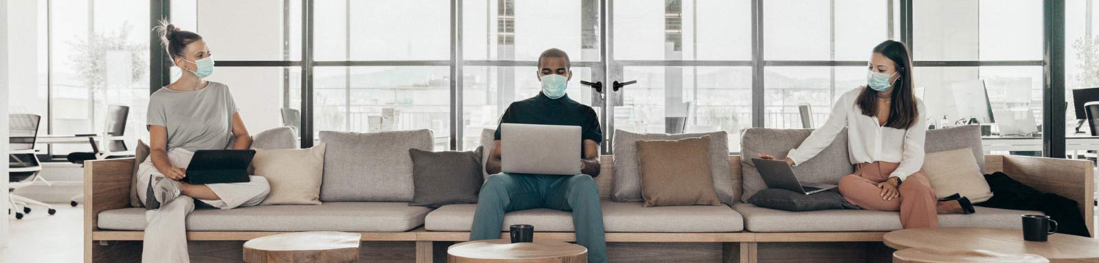 three masked people sitting apart on couch with laptops