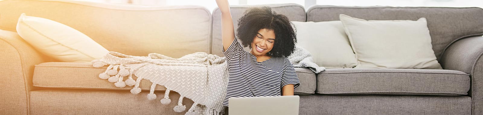 Woman smiling with fist in air sitting by her couch with a laptop