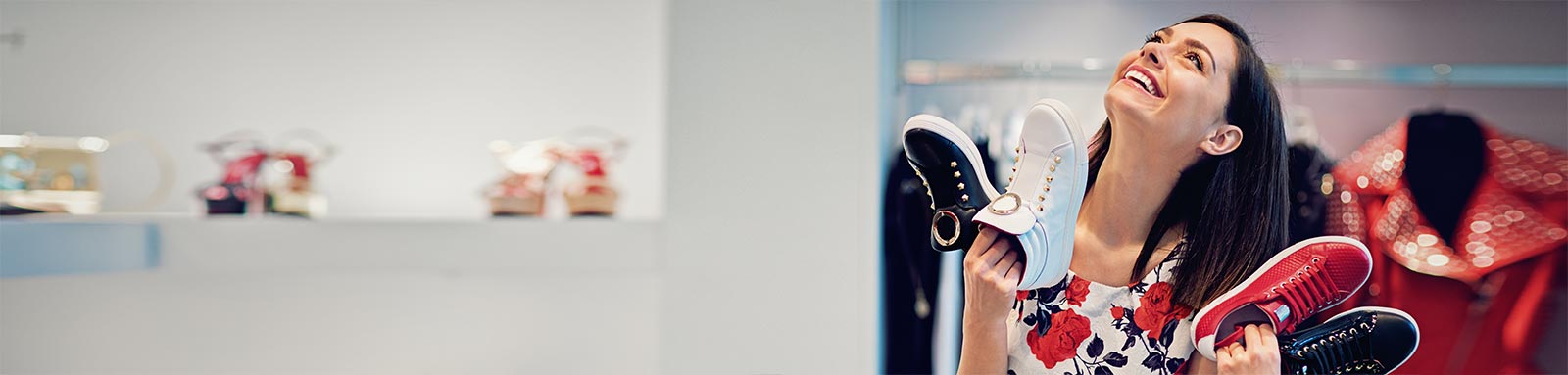 A woman in white floral frock holding shoes & smiling while shopping at shoe store