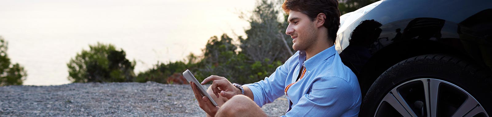 Young man on iphone sitting on the ground next to a black car